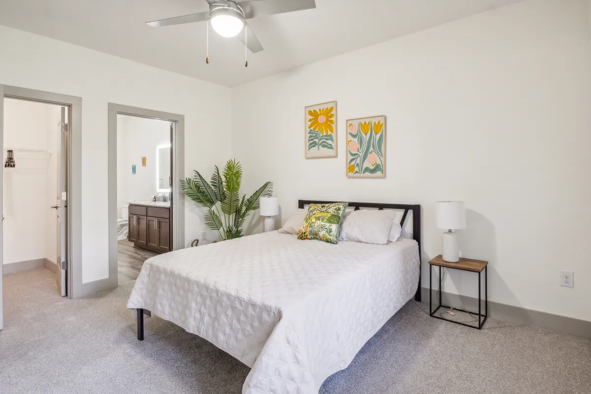 A cozy bedroom scene with a bed adorned with decorative pillows, stylish wall art, a potted plant, and a nightstand with a lamp. The room is bright and inviting, emphasizing comfort and a refreshing atmosphere.