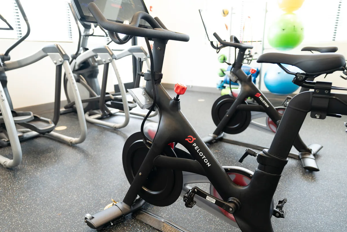 A bright, inviting fitness center featuring Peloton bikes, elliptical machines, and colorful exercise balls. The layout is spacious and well-lit, encouraging both solo and group workouts. Large windows (if present, implied by natural light) suggest a connection to the outdoors and the active lifestyle available to residents.