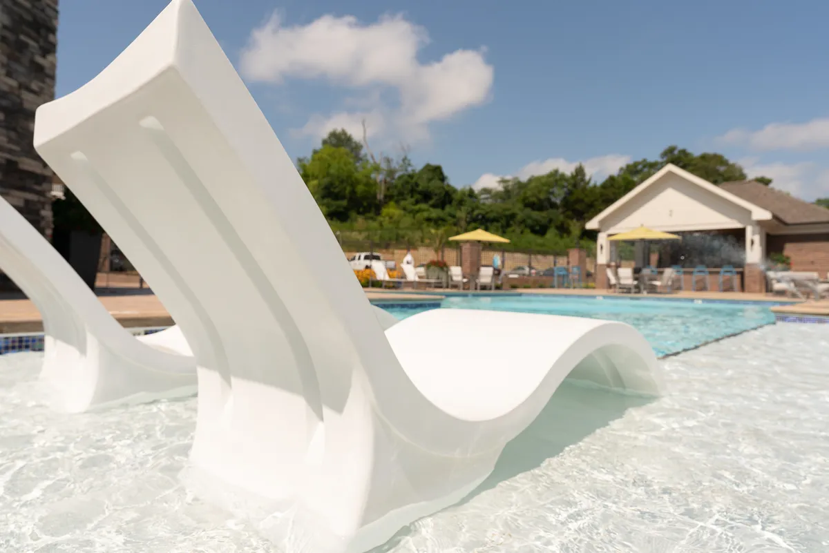 A sparkling community swimming pool is surrounded by lounge chairs, sun umbrellas, and lush greenery, with a stylish poolside building in the background. The setting is lively yet tranquil, ideal for socializing or relaxing on warm days.