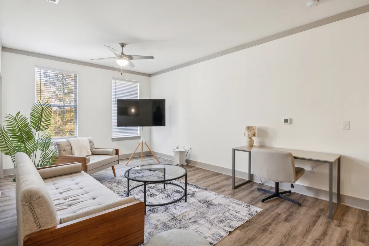 The image showcases a stylish, modern living space featuring a plush sofa, sleek coffee table, flat-screen TV, functional desk, and lush indoor plant. The arrangement is inviting, with natural light enhancing the cozy yet contemporary ambiance—ideal for relaxing, entertaining, or working remotely.