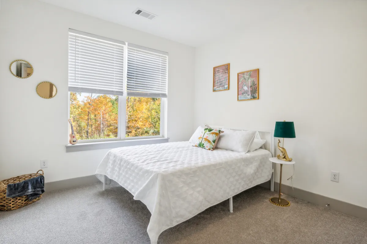 A beautifully lit bedroom with a bed dressed in a white quilted cover, a green lamp on the nightstand, decorative wall art, and a large window flooding the space with natural light. The scene exudes comfort and tranquility, with thoughtful touches that invite relaxation.