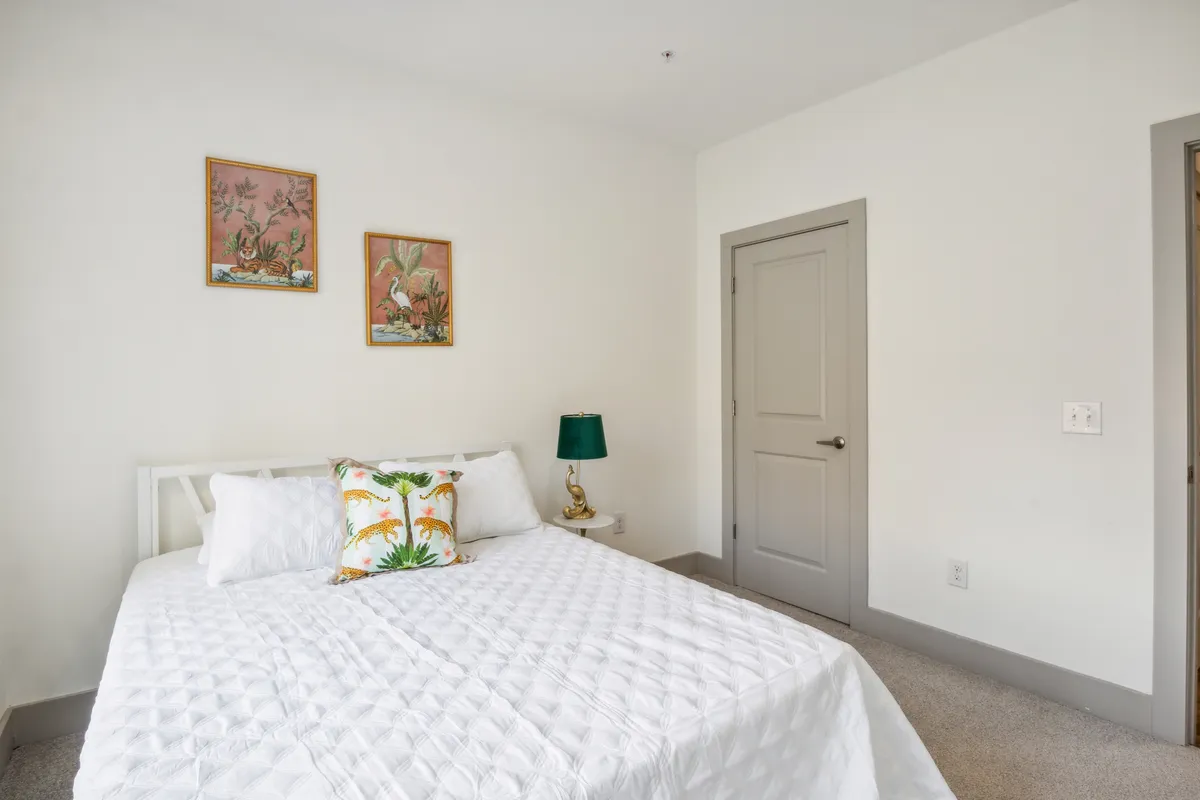This image features a well-lit bedroom with a neatly made bed draped in a white quilt, a green lamp on the nightstand, and framed artwork above. The sunlight streaming through the window creates a welcoming and comfortable ambiance, suggesting a restful space to unwind after exploring Knoxville.