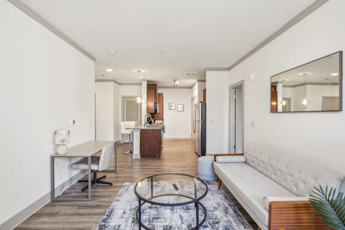 An open-concept living area featuring a modern sofa, a sleek coffee table, an adjacent dining area, and a view into the kitchen. The space is filled with light and styled with contemporary furnishings, offering both comfort and a welcoming atmosphere.