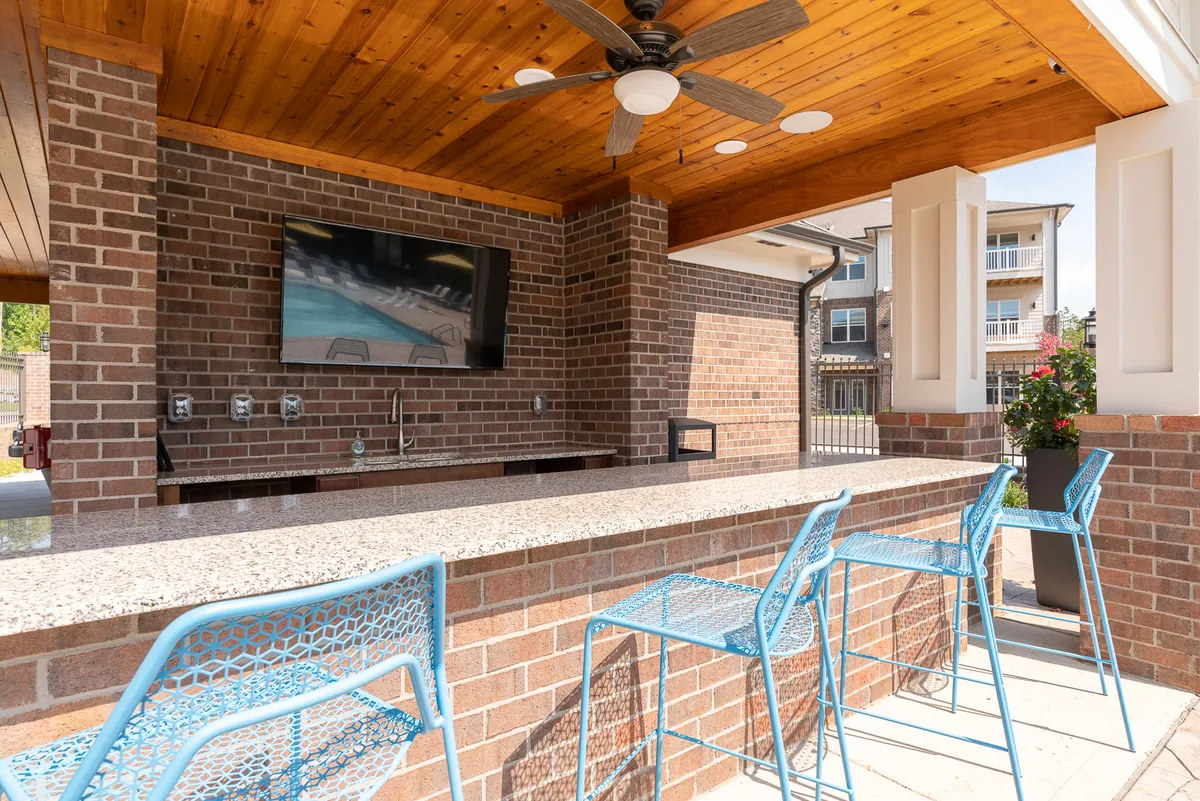 This exterior shot captures a contemporary outdoor lounge area, featuring a granite countertop with blue metal chairs, a wall-mounted TV, and a wooden ceiling. The space is designed for relaxing, socializing, and enjoying meals or entertainment outside.