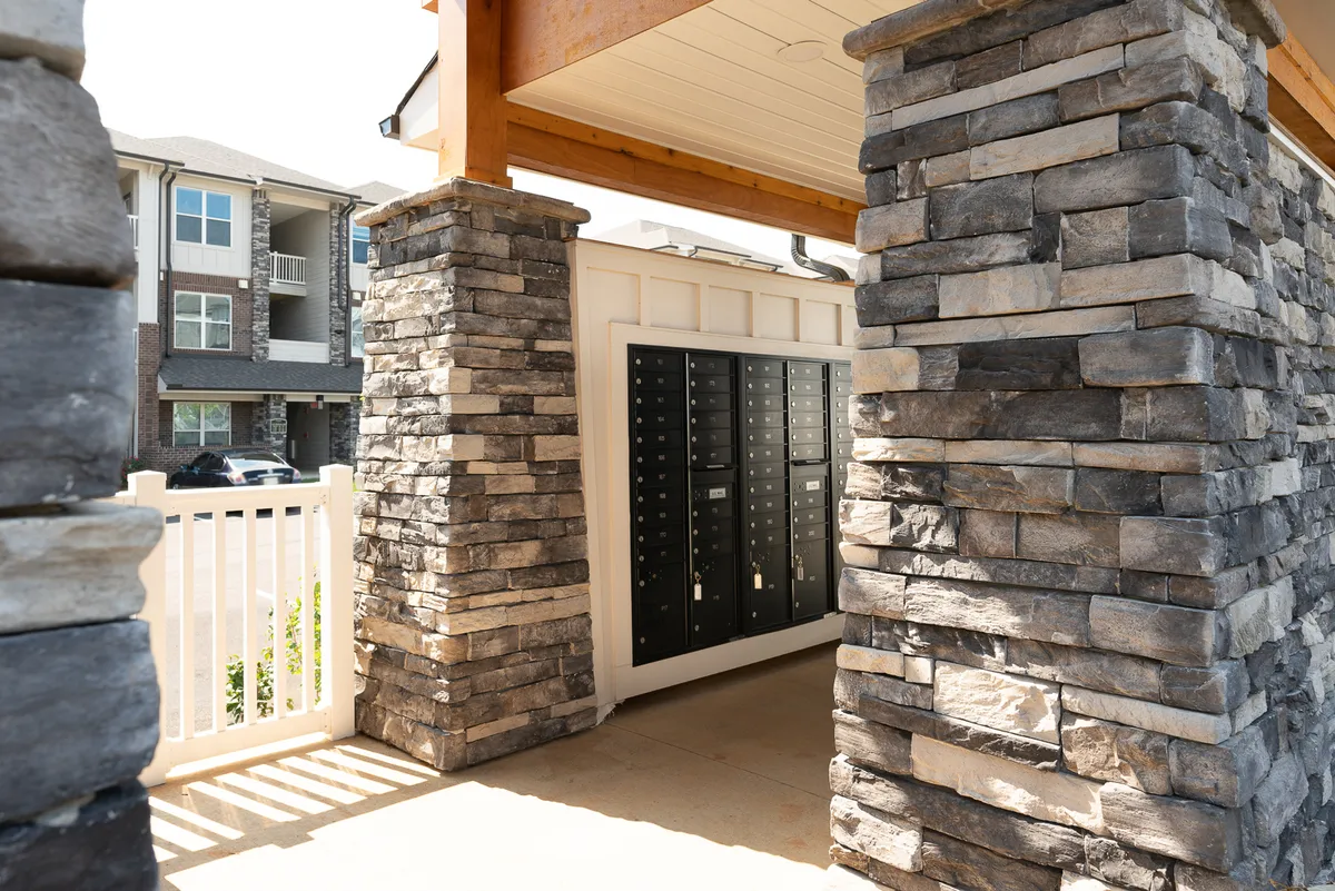 A welcoming exterior view of the apartment community's entrance, marked by stone columns, mailboxes, and a distinct entrance structure. Residents can be seen engaging in daily routines such as mail collection and friendly greetings.