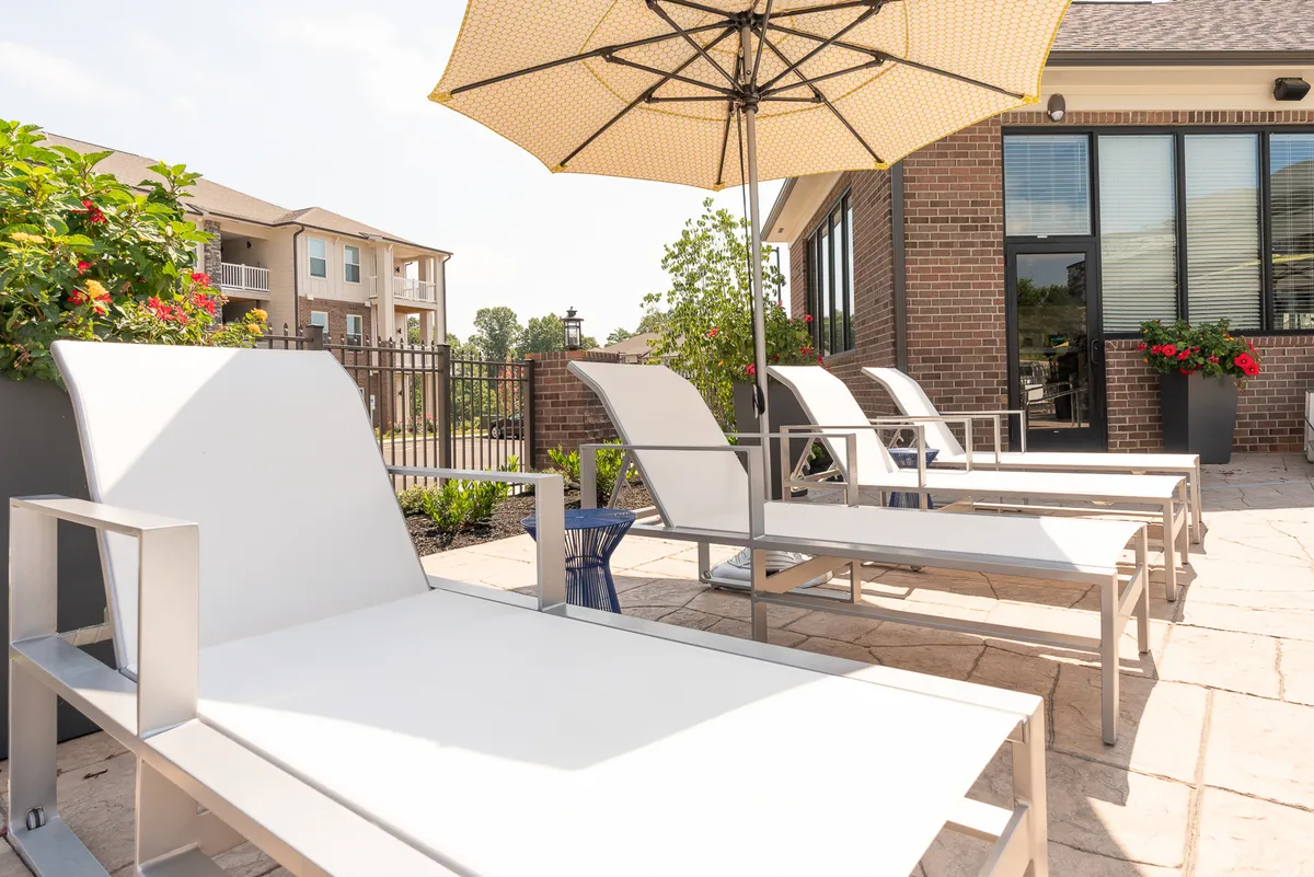 An inviting poolside area featuring lounge chairs, an umbrella, vibrant flower beds, and the backdrop of a brick building. The space exudes tranquility and offers options for sunbathing, reading, or casual gatherings.